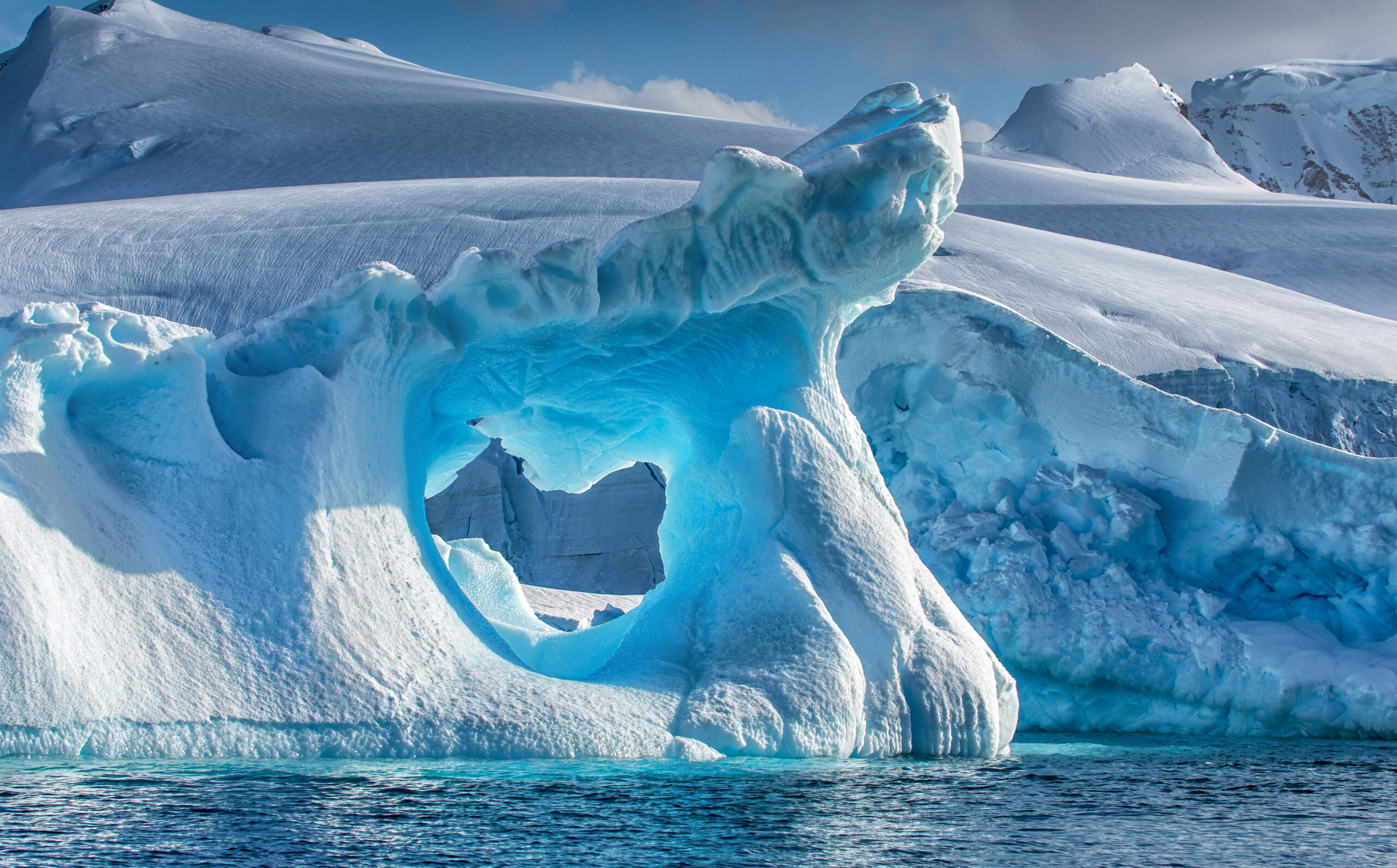 A dramatically shaped and weather eroded iceberg floating in Wilhemina Bay, Antarctica with a large hole in the centre glowing blue, and with a distant background of snow and ice covered mountains.
